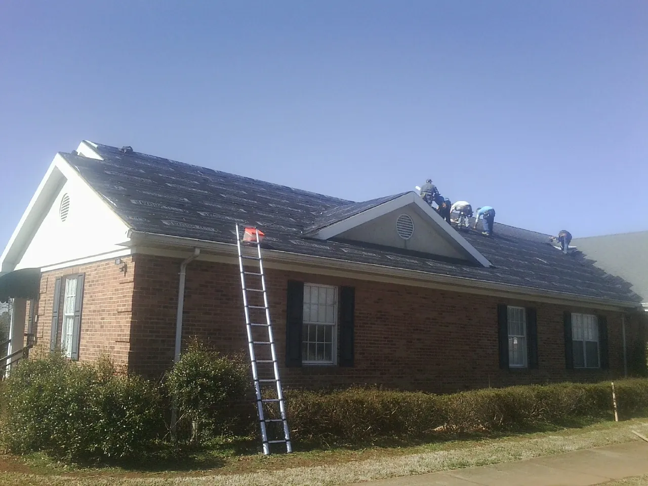 Crew tearing shingles off a brick ranch roof with a ladder set — Metal Roof Repair work by our crew in Black Creek